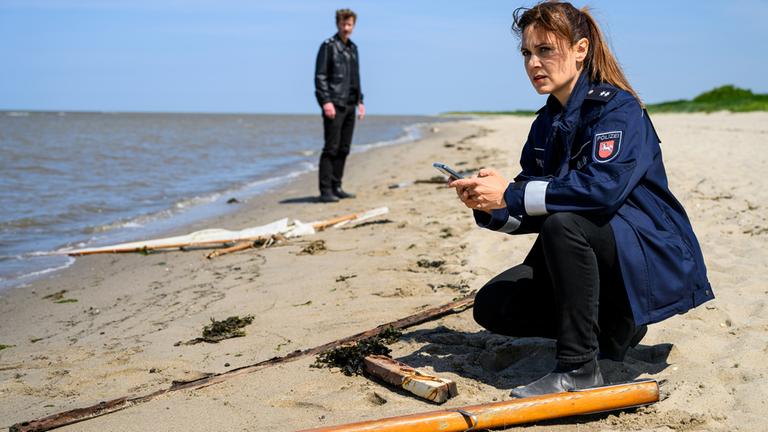 Süher Özlügül (Sophie Dal) kniet am Strand vor zerbrochenen und verstreuten Teilen eines Segelbootes. Im Hintergrund steht Henk Cassens (Maxim Mehmet).