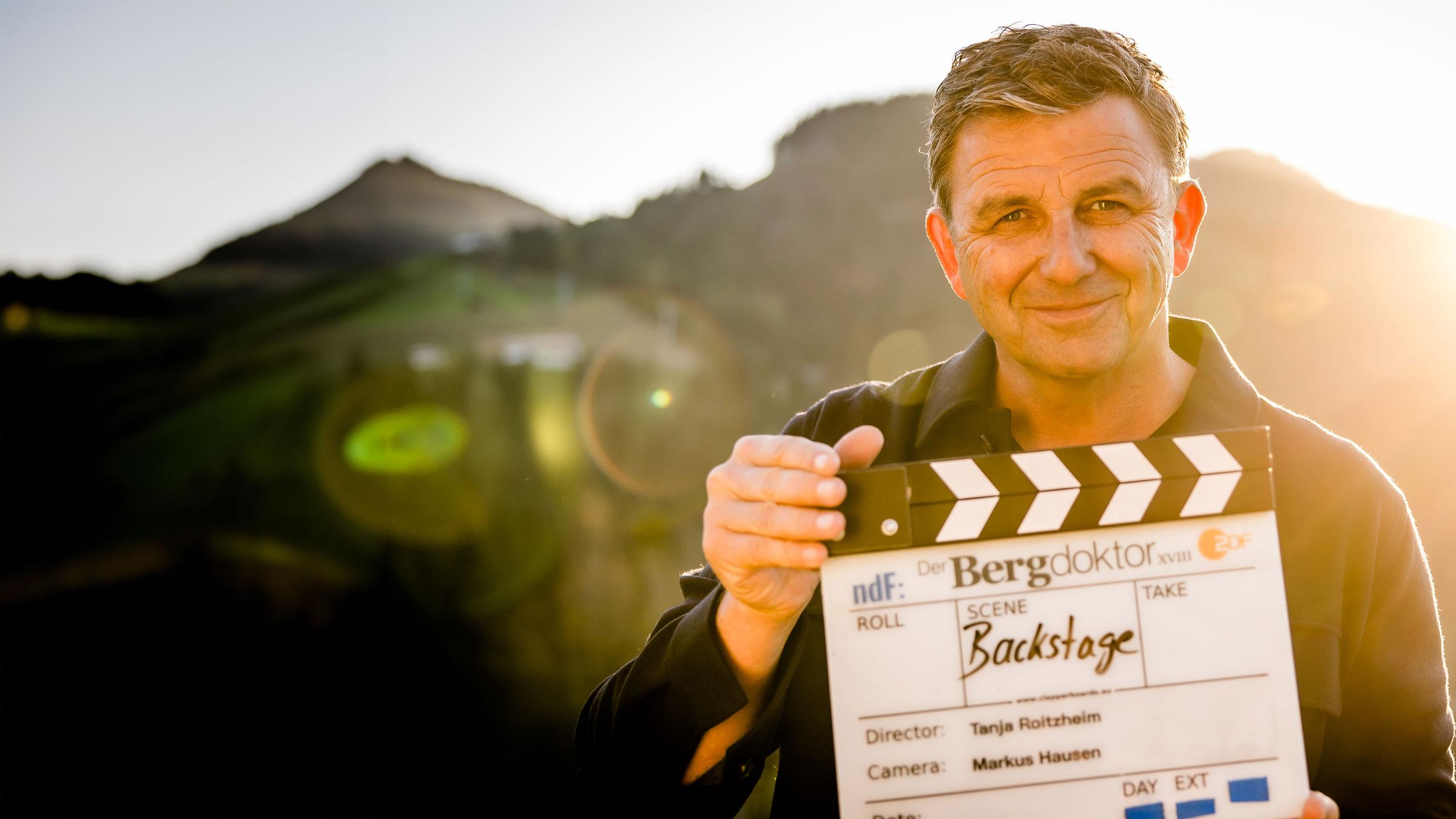 "Backstage - Der Bergdoktor: Am Set mit Hans Sigl": Schauspieler Hans Sigl steht bei Dreharbeiten in einer Berglandschaft. Er hält eine Filmklappe in der Hand und schaut lächelnd in die Kamera.