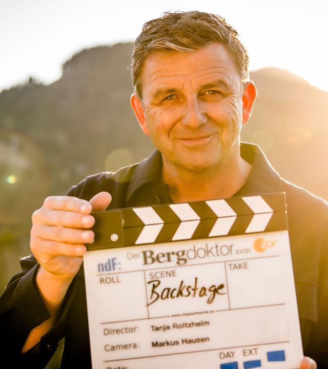 "Backstage - Der Bergdoktor: Am Set mit Hans Sigl": Schauspieler Hans Sigl steht bei Dreharbeiten in einer Berglandschaft. Er hält eine Filmklappe in der Hand und schaut lächelnd in die Kamera.