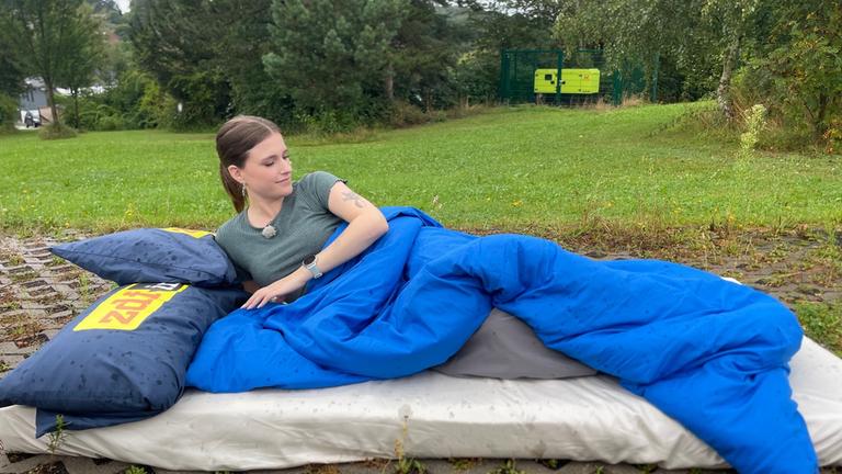 "Kann das was? Schlaf-Mythen im Check": Comedian Saskia Fröhlich liegt auf einer Matratze, die draußen auf dem Boden liegt, hinter ihr stehen Bäume an einer Wiese. Sie liegt seitlich unter einer blauen Bettdecke, am Kopfende liegt ein Kissen mit dem "ZDFinfo"-Logo. Sie schaut Richtung Bettende.