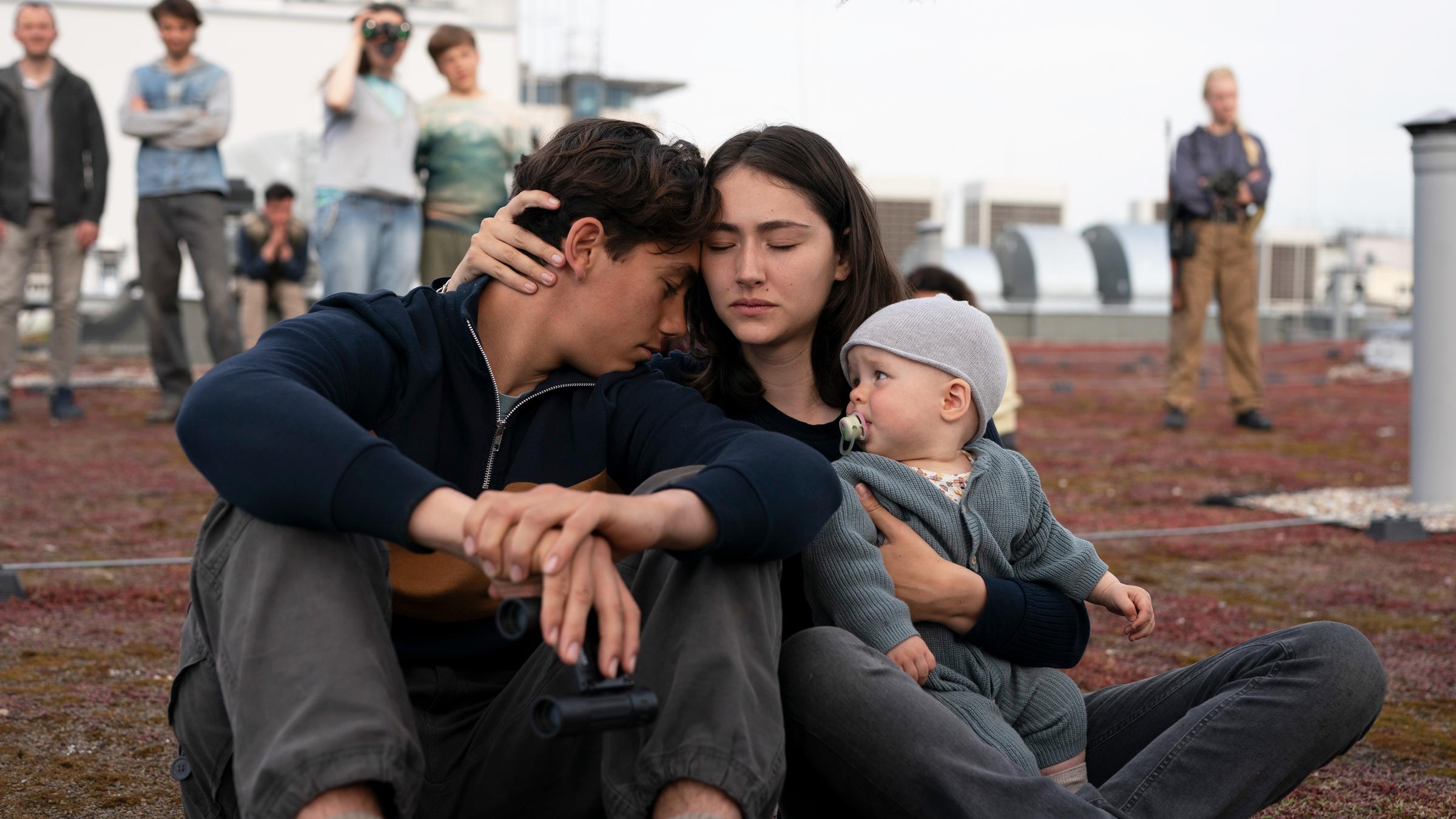 "Sløborn III – Eine neue Welt": Alex (Ron Renzenbrink) und Evelin (Emily Kusche) sitzen eng umschlungen mit Baby Mila (Lucia Wernze) im Arm auf einem mit Moos bewachsenen Flachdach. Im Hintergrund stehen mehrere Menschen.