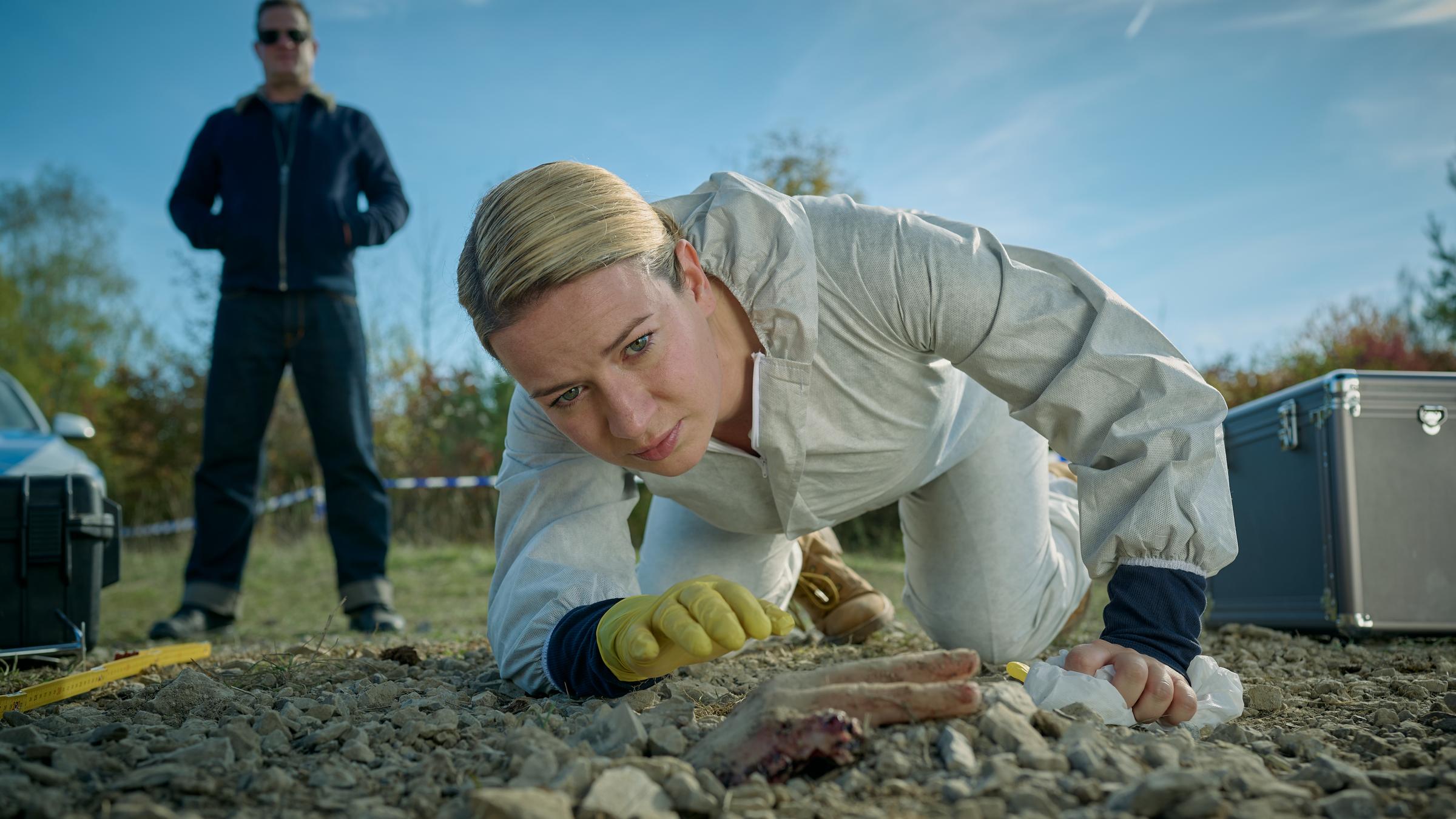 "Theresa Wolff - Dreck": Theresa Wolff (Nina Gummich) beugt sich über die freigelegte Hand einer Leiche. Sie trägt einen Schutzanzug und einen Gummihandschuh. Hinter ihr steht Bruno Lewandowski (Aurel Manthei) mit den Händen in seiner Jacke.