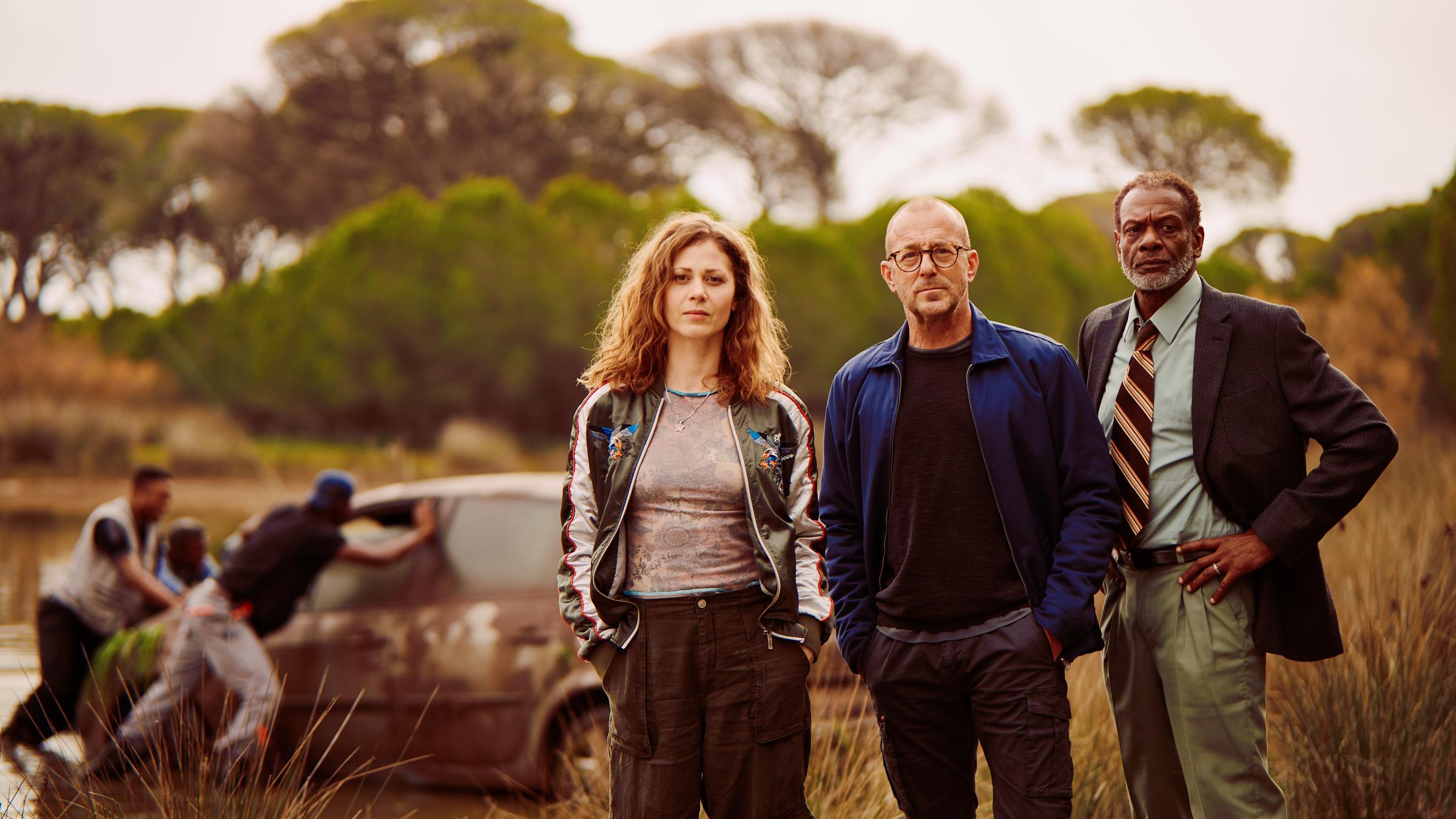 "Tod in Mombasa": Moritz Wagner (Heino Ferch), Caroline Jakobs (Katharina Schlothauer) und Kommissar Lomboto (Errol Trotman Harewood) stehen nebeneinander und schauen ernst in die selber Richtung. Hinter ihnen befindet sich ein See, aus welchem drei Männer gerade ein dreckiges Auto schieben. Hinter dem See stehen Pinien und andere Bäume.