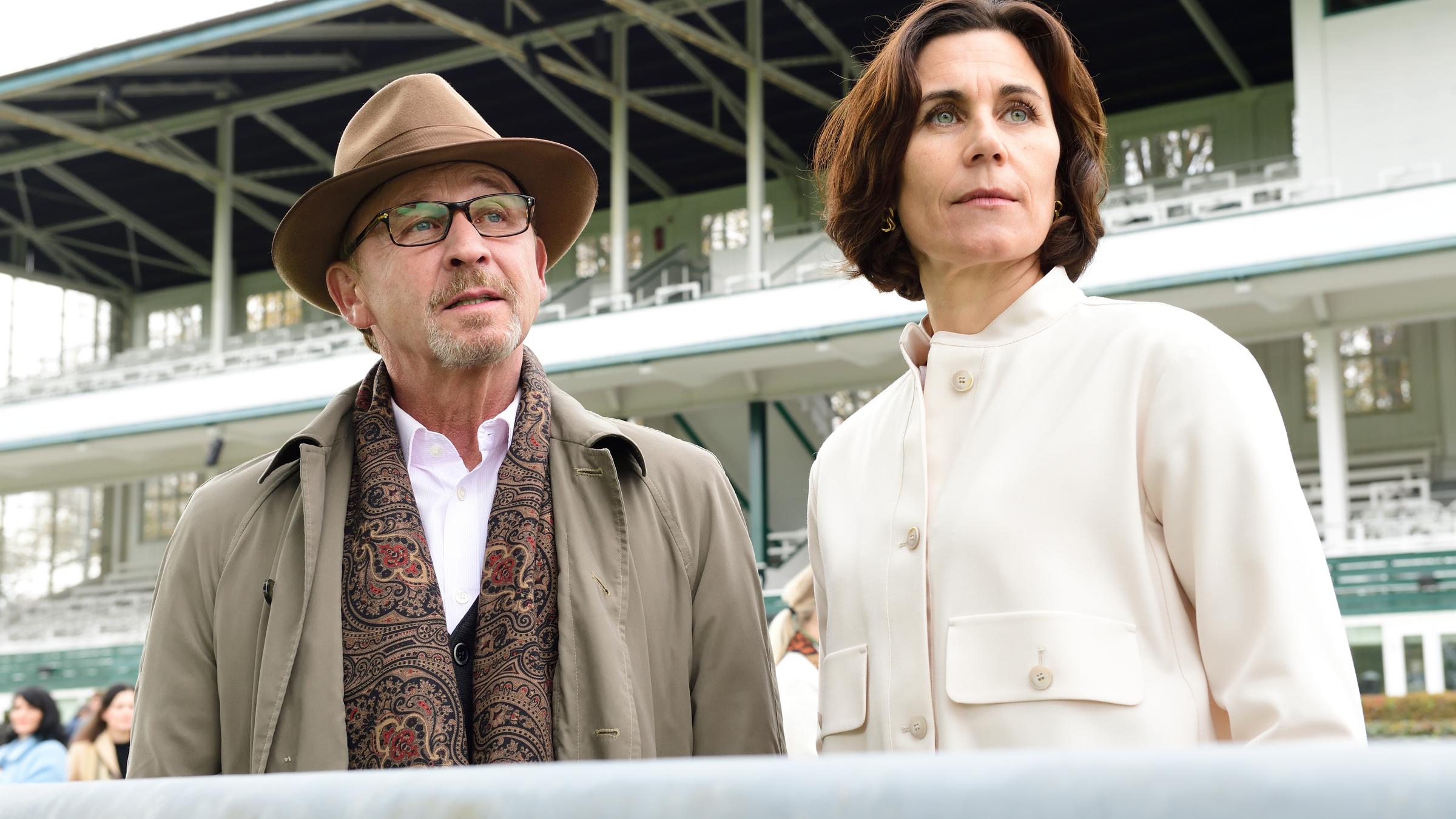 „München Mord - A saisonale G’schicht“: Ludwig Schaller (Alexander Held) und Kathrin Markgraf (Nina Kunzendorf) stehen nebeneinander an einem Schutzgeländer auf der Pferderennbahn und blicken nach vorne. Im Hintergrund befinden sich noch weitere Personen (Komparsen) auf einer Tribüne.