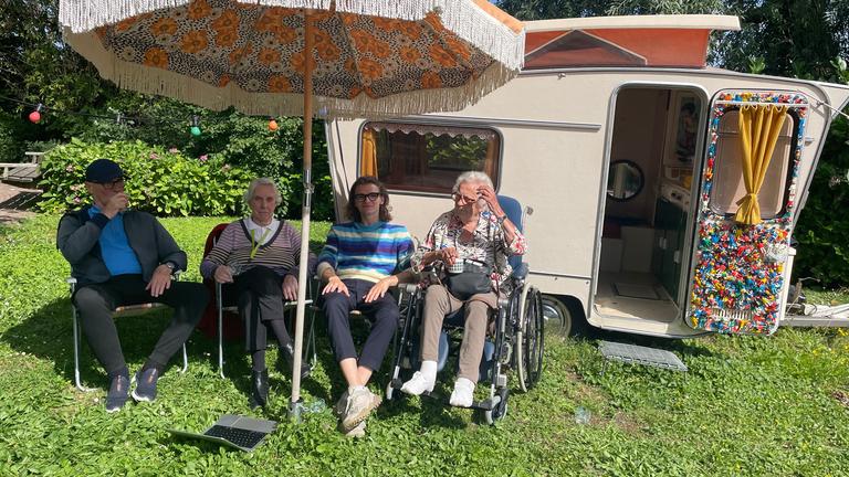 "plan b: Frischer Wind im Pflegeheim - Mehr Lebensqualität im Alter": Drei Pflegeheimbewohner sitzen mit einem jungen Mann im Garten unter einem Sonnenschirm vor einem kleinen Wohnwagen.