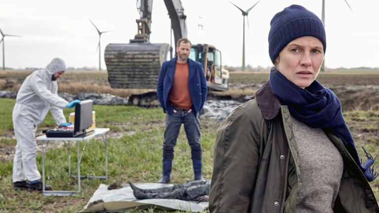 "Ostfriesenschwur": Kommissarin Ann Kathrin Klaasen (Picco von Groote) und ihr Kollege Frank Weller (Christian Erdmann) sind gemeinsam am Tatort. Klaasen steht im Vordergrund und blickt nachdenklich in die Ferne. Frank Weller steht hinter ihr. Vor ihm liegt eine Leiche. Ein Kollege (Komparse) von der Spurensicherung ist mit am Tatort und öffnet den Koffer mit den Utensilien für die Spurensicherung. Im Hintergrund stehen Windkrafträder und davor ein Bagger.