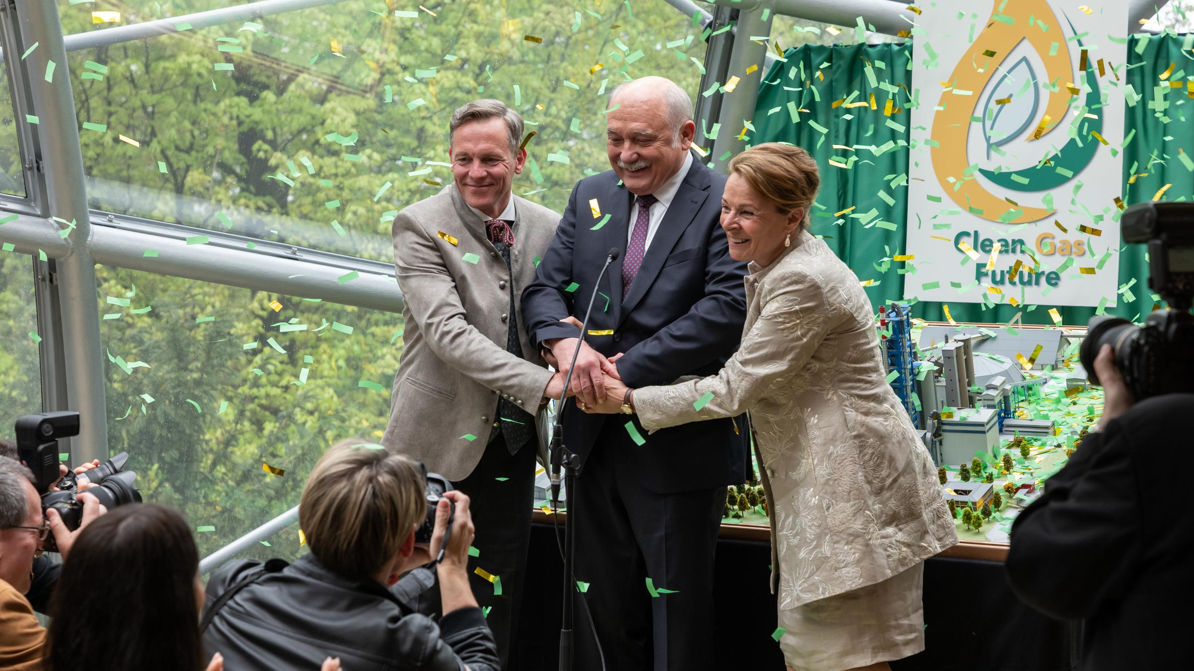 "Die Toten von Salzburg – Süßes Gift": Josef Wittmann (Johannes Zirner), Gustav Nussbaumer (Karl Fischer) und Susanne Zirner (Susanne Czepl) stehen zusammen auf einem Podest vor Journalisten (Komparsen). Sie haben ihre Hände in der Mitte aufeinander gelegt und es regnet Konfetti auf sie. Im Hintergrund hängt ein Plakat mit der Aufschrift "Clean Future".