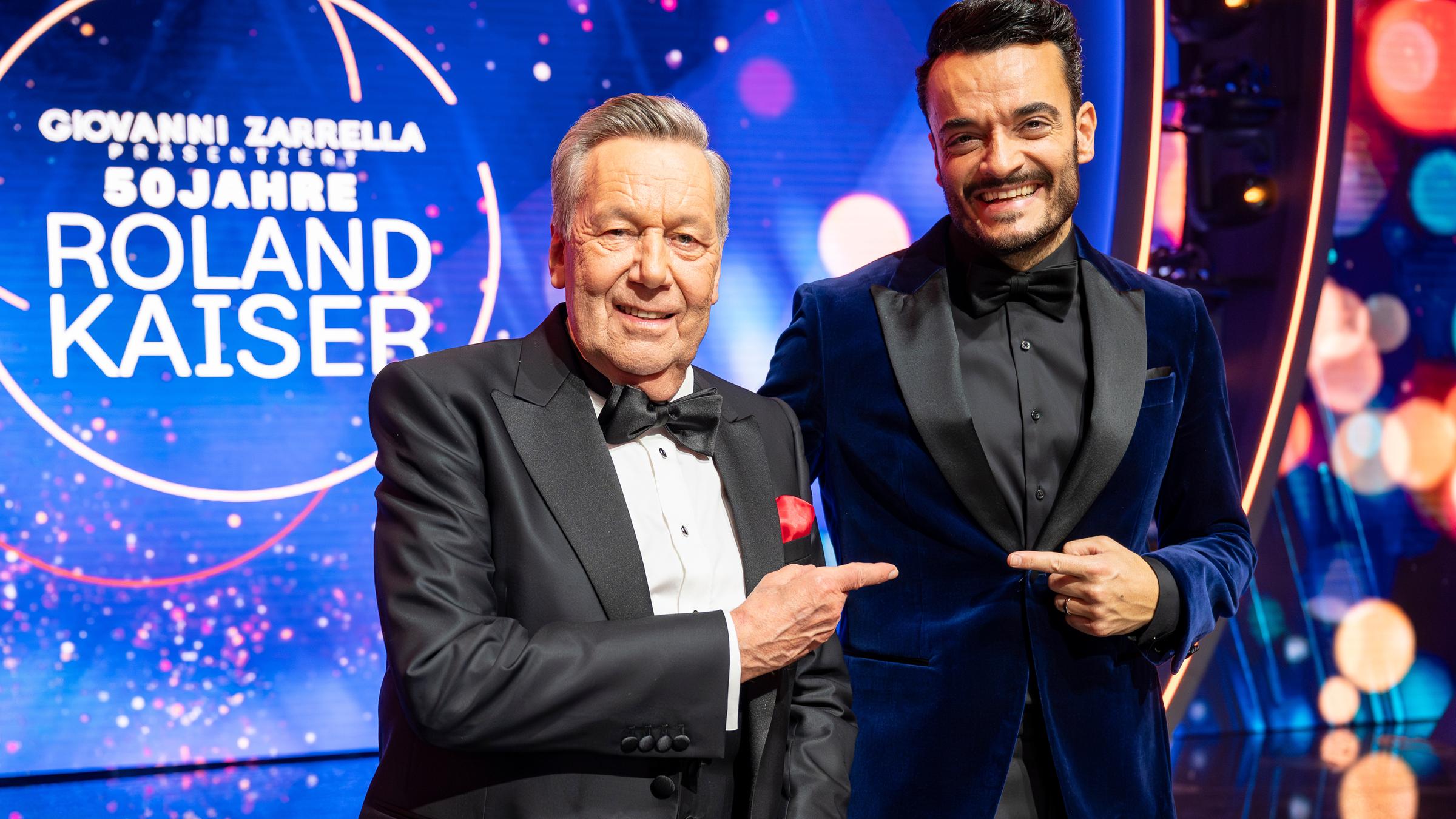 "Giovanni Zarrella präsentiert: 50 Jahre Roland Kaiser": Roland Kaiser und Giovanni Zarrella stehen nebeneinander vor einer LED-Wand, auf der das Sendungslogo der Show zu sehen ist. Beide sind elegant im dunklen Smoking gekleidet und lachen in die Kamera.