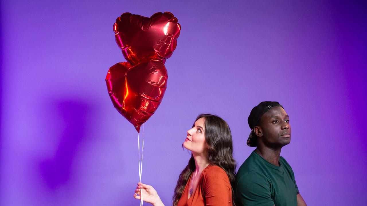 Zwischen Liebe, Lust und Herzschmerz (ZDF-Programmakzent): Mia und Dennis stehen Rücken an Rücken vor einem lila Hintergrund. Mia hält in der rechten Hand zwei rote Luftballons in Herzform