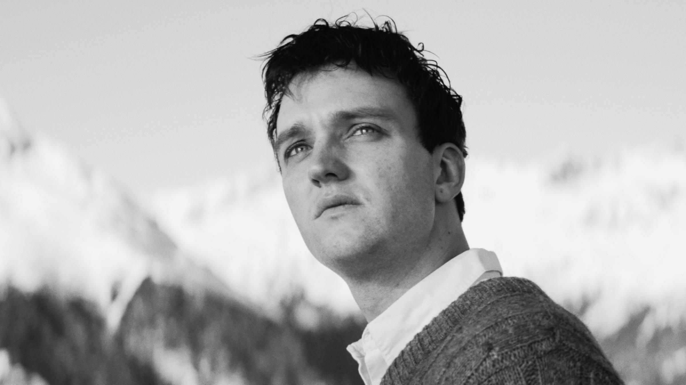 "Die Theorie von Allem" (SW-Foto): Johannes Leinert (Jan Bülow) blickt mit linksgewandtem Kopf in die Kamera. Im Hintergrund sieht man verschwommen schneebedeckte Berge.