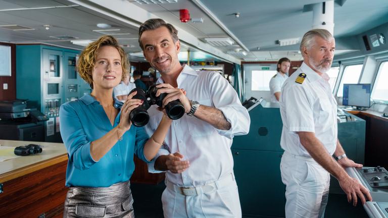 "Das Traumschiff - Nusantara": Auf der Schiffsbrücke: Kapitän Max Parger (Florian Silbereisen) übergibt  Veronika Bruckner (Wanda Perdelwitz) ein Fernglas. Beide blicken lächelnd in die Ferne. Im Hintergrund steht Staff-Kapitän Martin Grimm (Daniel Morgenroth) und beobachtet die beiden.
