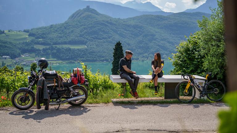 "Horst Lichters Traumrouten - Von Meran nach Venedig": Horst Lichter und Dorothea Wierer sitzen auf einer Bank. Neben Horst Lichter steht sein Motorrad und neben Dorothea Wierer ihr Fahrrad. Im Hintergrund liegt der Kalterer See und Berge.