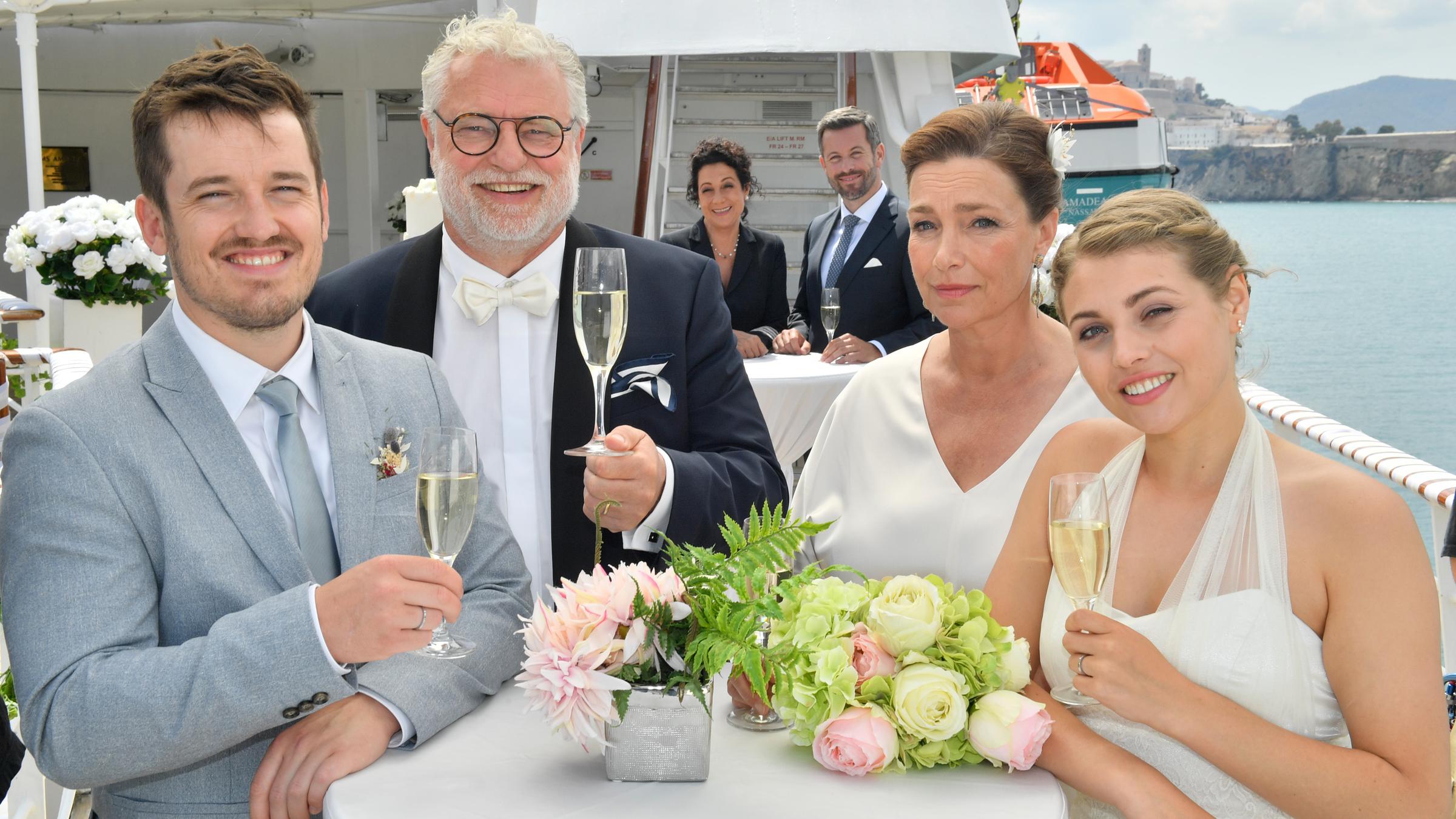 "Kreuzfahrt ins Glück - Hochzeitsreise nach Korsika": Auf dem festlich geschmückten Deck des Schiffs: Die Brautpaare Sabine (Aglaia Szyszkowitz) und Jean Rossi (Filip Peeters) sowie Ina Rossi (Nicola-Rabea Langrzik) und Tino Möller (Felix Kreutzer) stehen an einem Stehtisch und lächeln in die Kamera. Bis auf Sabine Rossi halten alle gefüllte Sektgläser in der Hand. Im Hintergrund stehen Hannah Liebhold (Barbara Wussow) und Tom Cramer (Jan Hartmann), ebenfalls an einem Stehtisch, und beobachten die Paare strahlend.