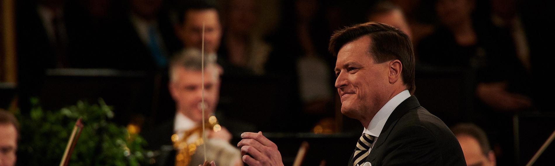 "Neujahrskonzert der Wiener Philharmoniker 2024": Der Dirigent Christian Thielemann steht mit erhobenen Armen vor einem kleinen Teil der spielenden Orchestermitglieder. Er lächelt dem Orchester zu und hält einen Taktstock in der rechten Hand. Thielemann trägt einen eleganten schwarzen Anzug mit einem weißen Hemd und einer gestreiften Krawatte.