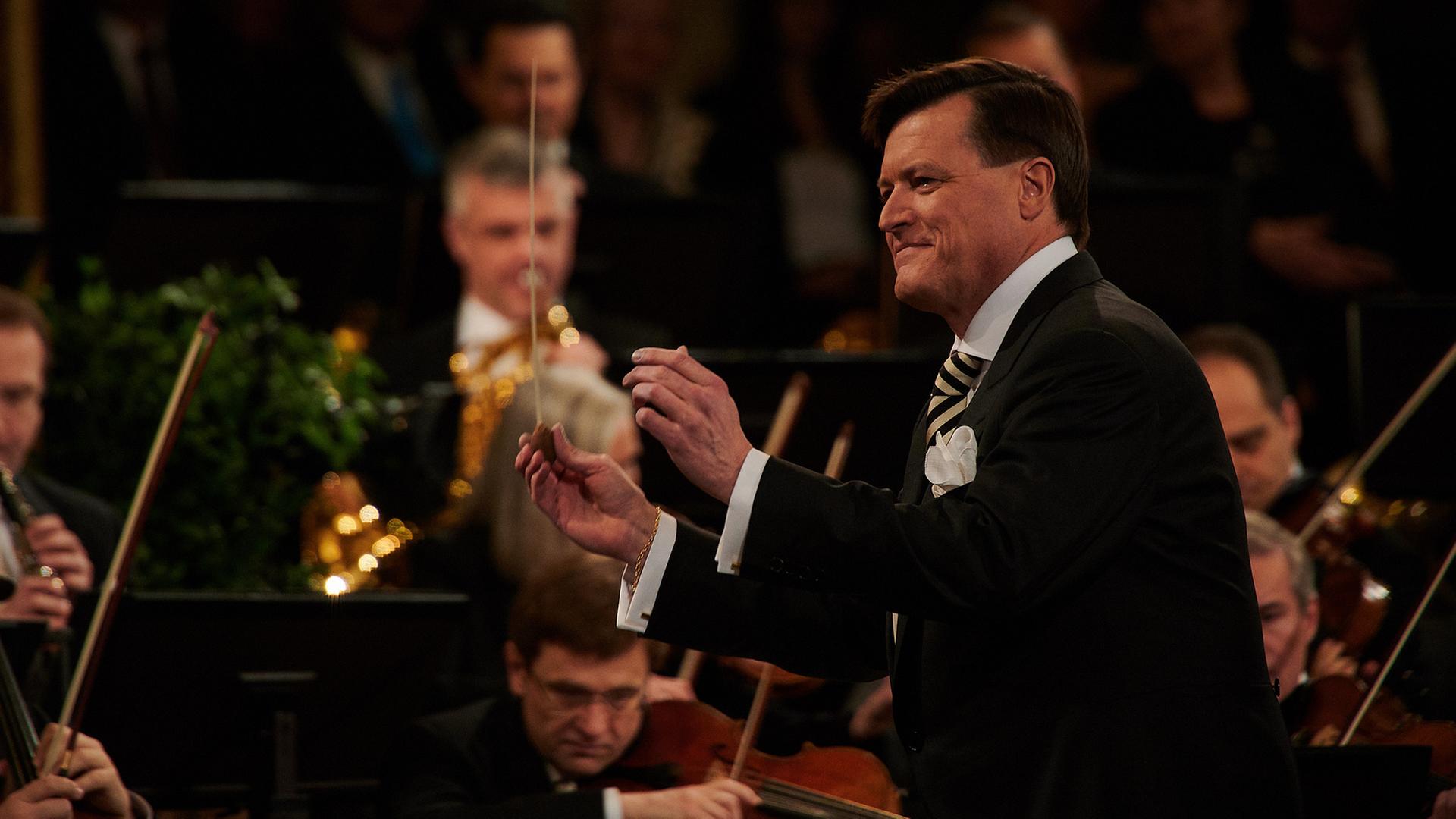 "Neujahrskonzert der Wiener Philharmoniker 2024": Der Dirigent Christian Thielemann steht mit erhobenen Armen vor einem kleinen Teil der spielenden Orchestermitglieder. Er lächelt dem Orchester zu und hält einen Taktstock in der rechten Hand. Thielemann trägt einen eleganten schwarzen Anzug mit einem weißen Hemd und einer gestreiften Krawatte.