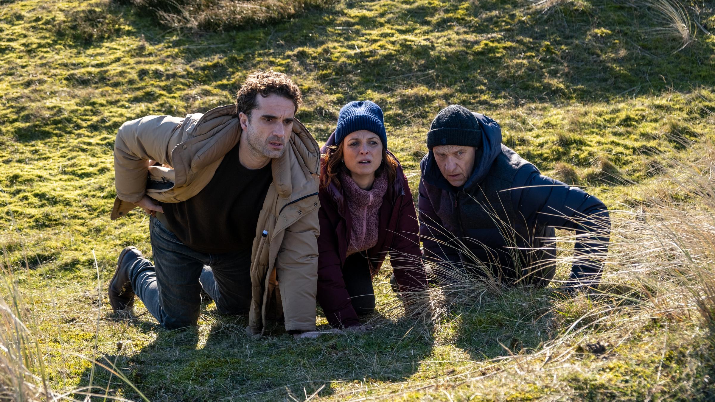 Die drei Kommissare Hinnerk Feldmann (Oliver Wnuk), Ina Behrendsen (Julia Brendler) und Carl Sievers (Peter Heinrich Brix) blicken angespannt, auf die Knie gestützt und geduckt, über den Kamm einer Düne. Neben ihnen stehen hohe Gräser.
