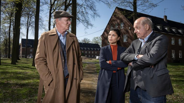 "Wilsberg - Ein Detektiv und Gentleman": Wilsberg (Leonard Lansink) und Tessa Tilker (Patricia Meeden) stehen mit verschränkten Armen neben John Cross (August Zirner) und schauen ihn kritisch an. John Cross schaut Wilsberg und Tessa ebenfalls an, seine Hände in den Manteltaschen vergraben. Er ist gekleidet wie ein britischer Gentleman, mit Schiebermütze und dreiteiligem Anzug. Im Hintergrund steht ein alte Kasernengebäude zwischen hohen Bäumen.