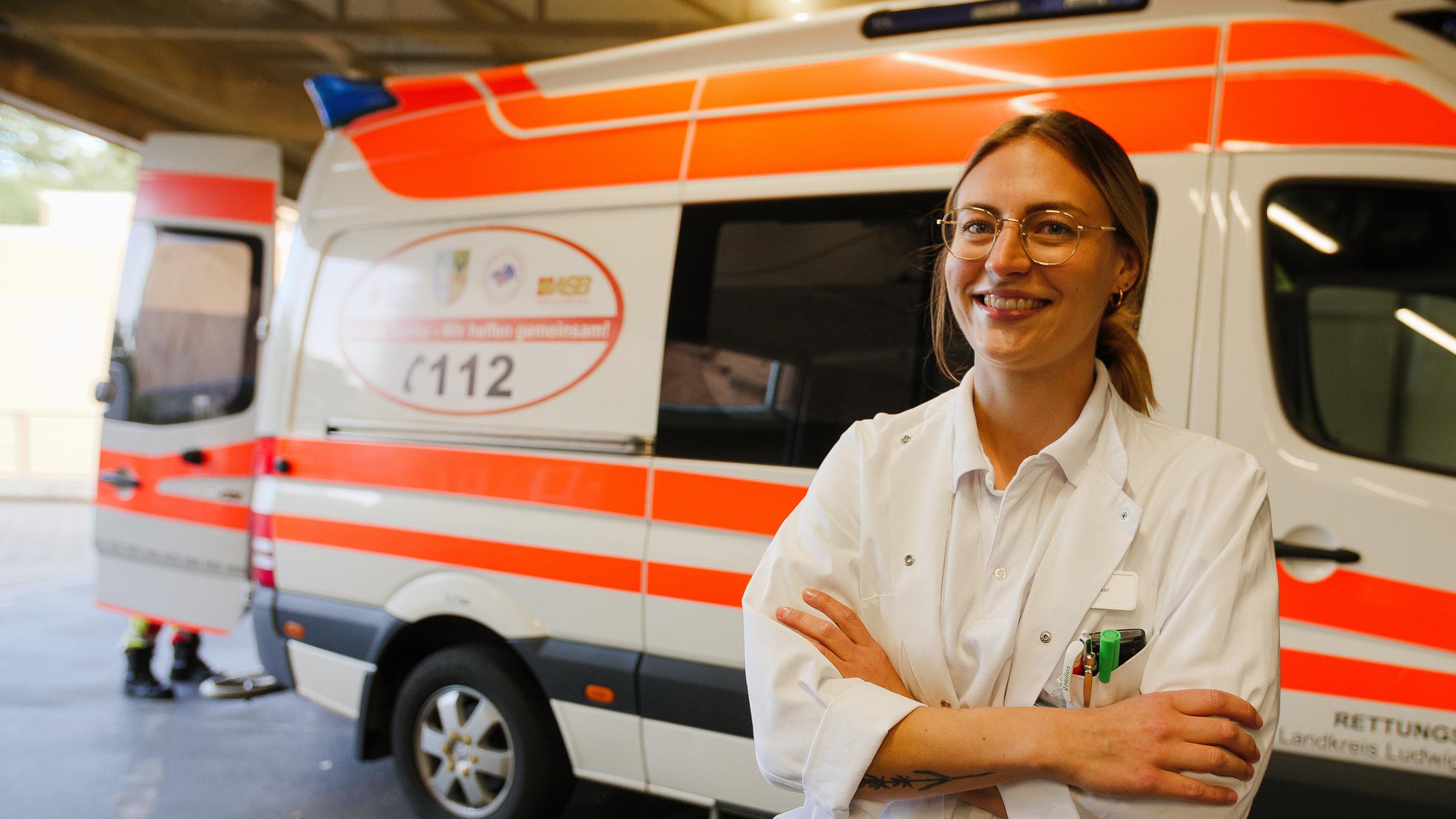 "37 Grad: Dienste, Druck und Dauerstress - Junge Docs in der Klinik": Assistenzärztin Jule (26) steht vor einem Rettungswagen und lächelt in die Kamera. Sie trägt einen weißen Arztkittel, eine Brille und ihre blonden langen Haare zu einem Zopf.