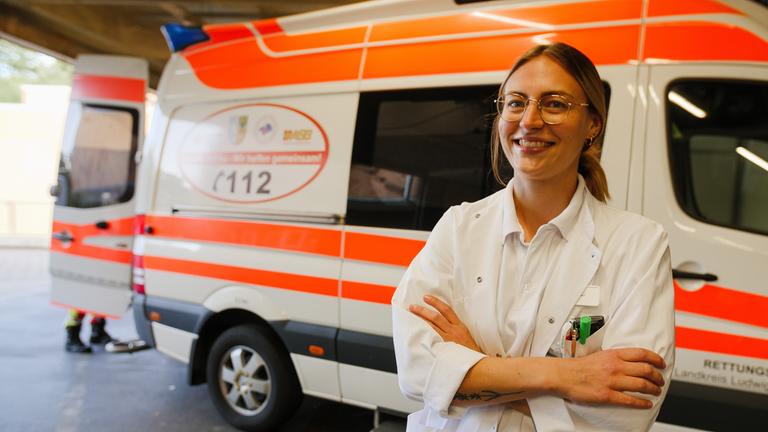 "37 Grad: Dienste, Druck und Dauerstress - Junge Docs in der Klinik": Assistenzärztin Jule (26) steht vor einem Rettungswagen und lächelt in die Kamera. Sie trägt einen weißen Arztkittel, eine Brille und ihre blonden langen Haare zu einem Zopf.