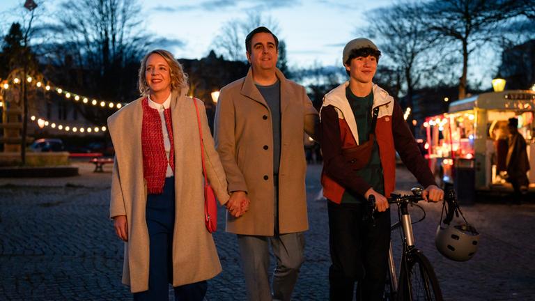"Alles gelogen": Am Abend: Vera (Katrin Wichmann), Hajo (Bastian Pastewka) und Marvin Siewers (Arthur Gropp) laufen lächelnd nebeneinander über einen mit Lichterketten geschmückten Platz. Marvin  schieb ein Fahrrad. Im Hintergrund steht ein beleuchteter Foodtruck.