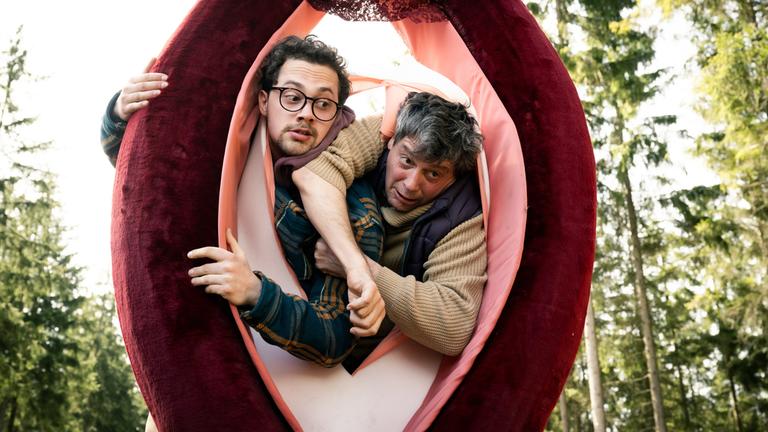 "Überväter": Luca (Anselm Bresgott) und Mathi (Fritz Karl) versuchen beide gleichzeitig durch eine überlebensgroße Vulva zu klettern und blockieren sich gegenseitig.