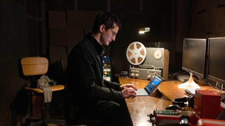 "Black Box - Gefährliche Wahrheit": Halbnahe: Mathieu (Pierre Niney) sitzt an einem Schreibtisch mit professioneller Audiotechnik und schaut konzentriert auf einen Tablet-PC mit Tastatur.