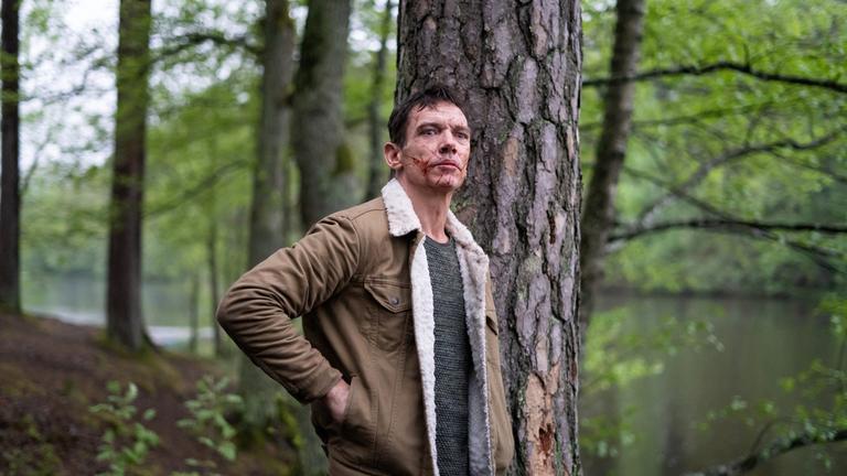 "The Good Neighbor": Robert (Jonathan Rhys Meyers) lehnt mit blutigen Narben im Gesicht und verstörendem Blick an einem Baum nahe eines Waldsees.