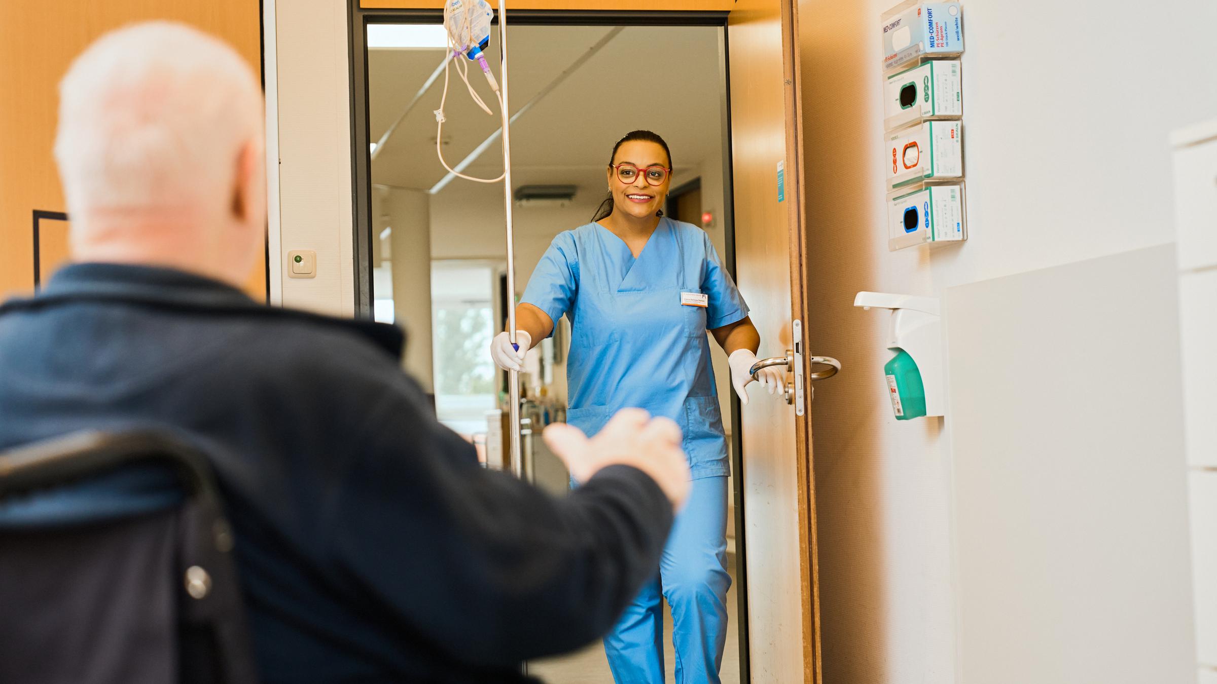 "37°: Arbeitskräfte weltweit gesucht!": Im Krankenhaus: Elaine, die Pflegekraft steht lächelnd in der offenen Tür. Ein älterer Mann im sitzt, mit dem Rücken zur Kamera, im Rollstuhl in einem Zimmer.