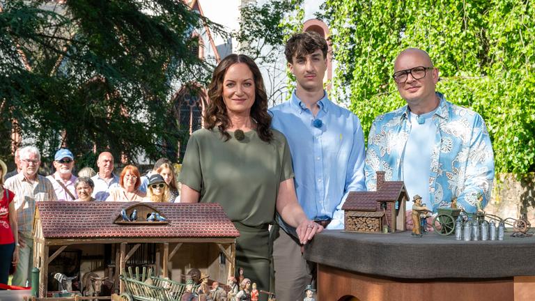 "Bares für Rares - Deutschlands größte Trödelshow": Natalia Wörner, Jacob Lee Seeliger und Sven Deutschmanek stehen hinter dem Expertisentisch im Freien. Auf dem Tisch ist ein Miniatur-Bauernhof aufgebaut. Alle schauen lächelnd in die Kamera. Im Hintergrund steht das Publikum.