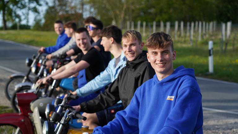 "ZDF.reportage: Jung im Osten – Wie wir wirklich leben": Jason aus Flurstedt und andere Jugendlichen sitzen auf Simson-Mopeds und lächeln in die Kamera. Im Hintergrund verläuft eine schmale Landstraße an der Bäume stehen.