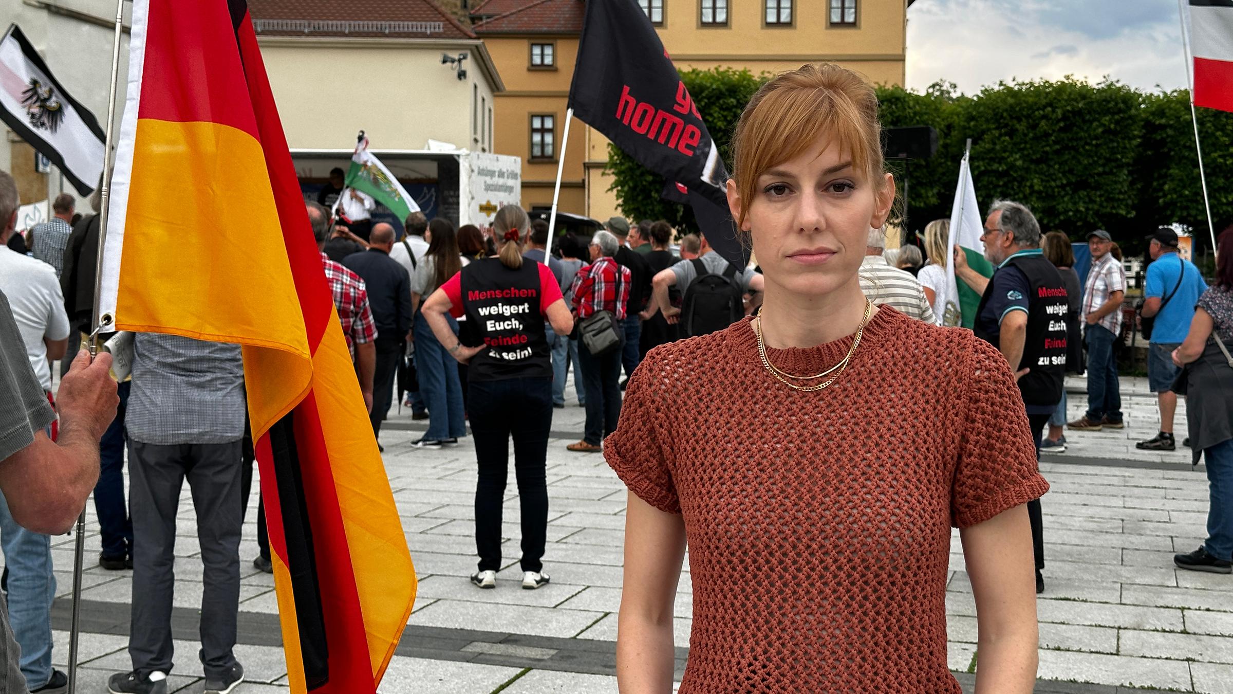 "Sachsen, warum bist du so gespalten?": Eva Schulz bei einer Demo in Sachsen: Sie steht auf einem Platz und schaut in die Kamera. Hinter ihr stehen viele Menschen, eine Person hält eine Deutschlandfahne, auf einer anderen steht "Ami go home".