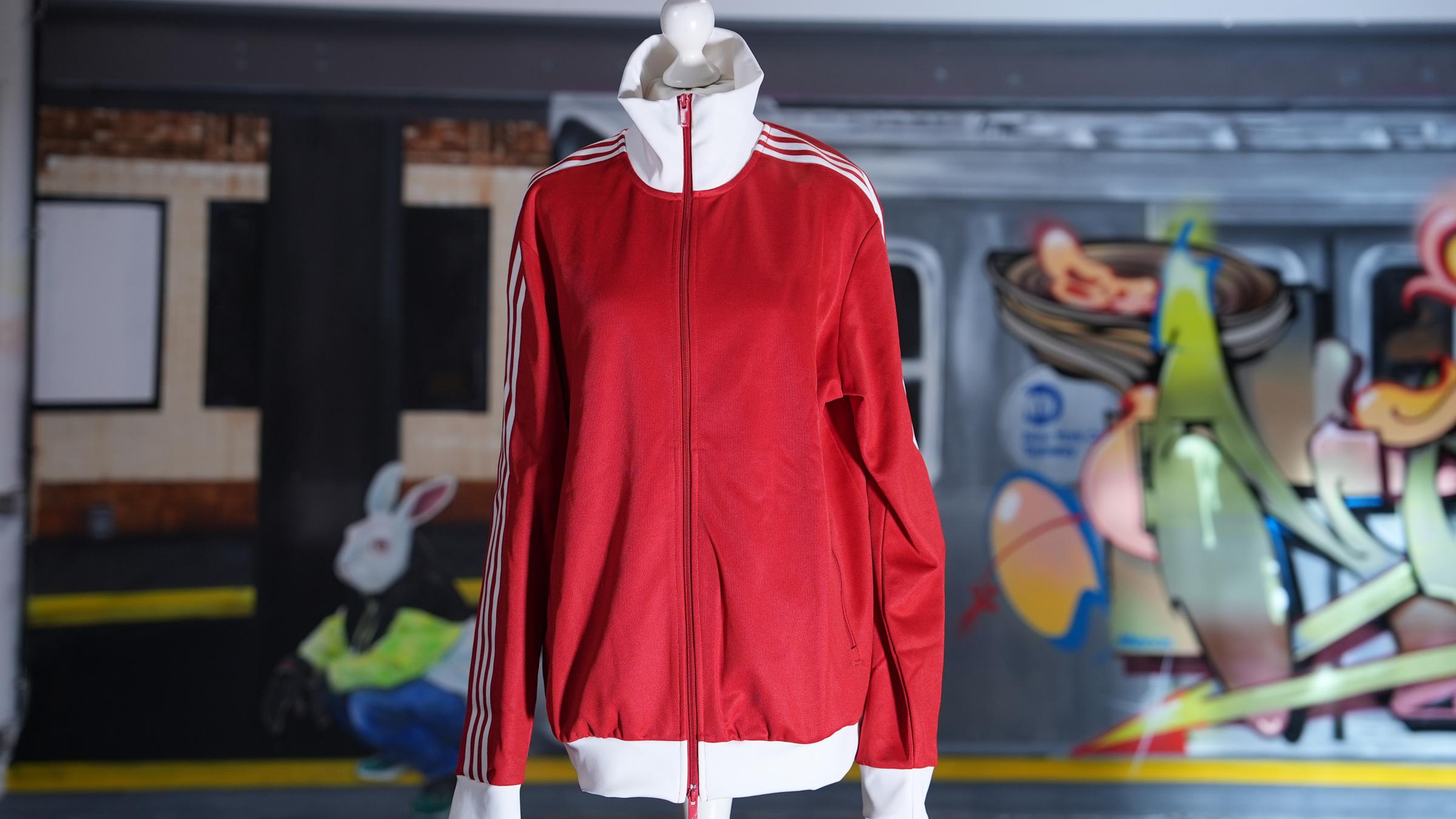 "planet e.: Luxus, Glamour, schöner Schein - Fast Fashion Jogginganzüge": Eine rote Trainingsjacke ohne Logos hängt auf einer Schneiderpuppe. Im Hintergrund steht ein mit Graffitis verzierter Waggon.