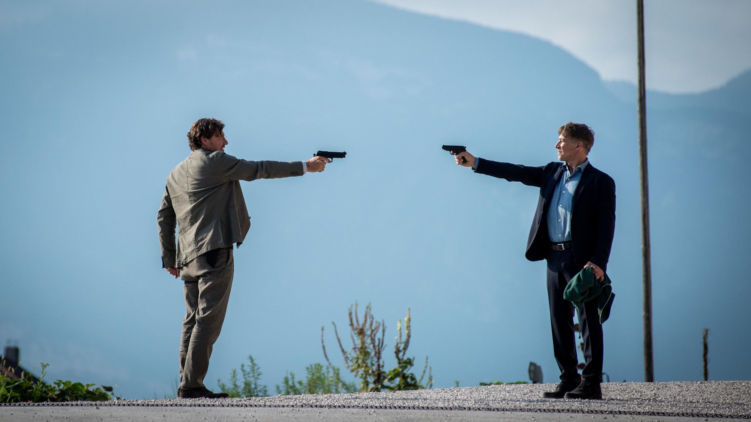 "Der Gejagte - Im Netz der Camorra": Adrin Erlacher (Harald Windisch) und Matteo DeCanin (Tobias Moretti) stehen sich auf einem Hügel gegenüber. Beide haben eine Pistole auf den jeweils anderen gerichtet, DeCanin hat in seiner anderen Hand einen grünen Pullover. Im Hintergrund lassen sich Berge erahnen.