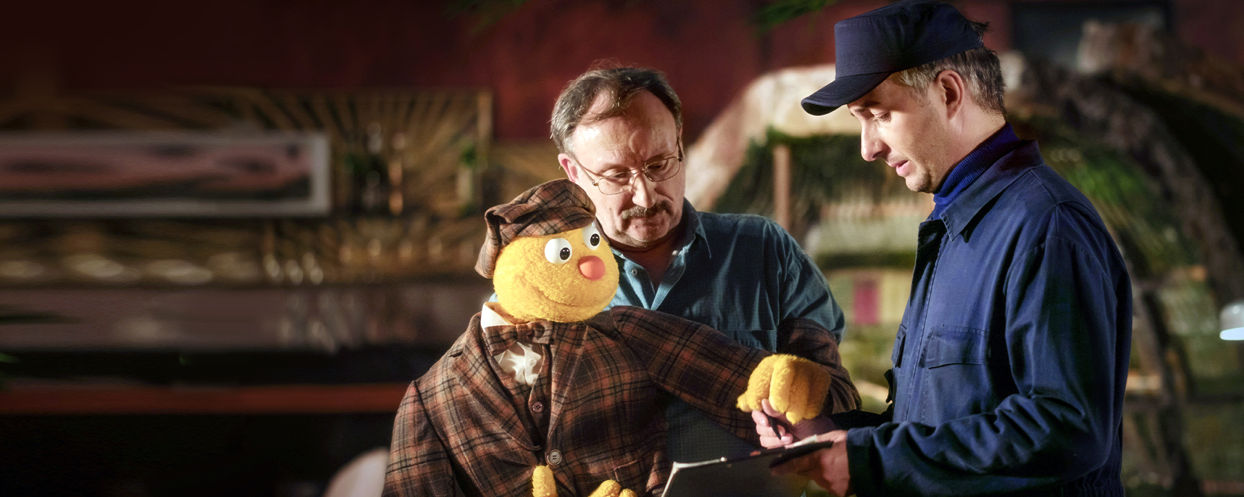 "Hallo Spencer - Der Film:" Die Handpuppe Spencer und Puppenspieler Jakob Sesam (Rainer Bock) unterschreiben einen Lieferschein, den ein Mann in blauer Arbeitskleidung (Jan Böhmermann) ihnen hinhält.