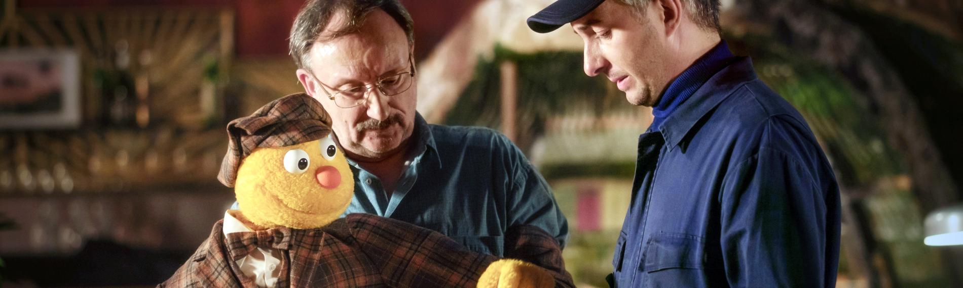 "Hallo Spencer - Der Film:" Die Handpuppe Spencer und Puppenspieler Jakob Sesam (Rainer Bock) unterschreiben einen Lieferschein, den ein Mann in blauer Arbeitskleidung (Jan Böhmermann) ihnen hinhält.