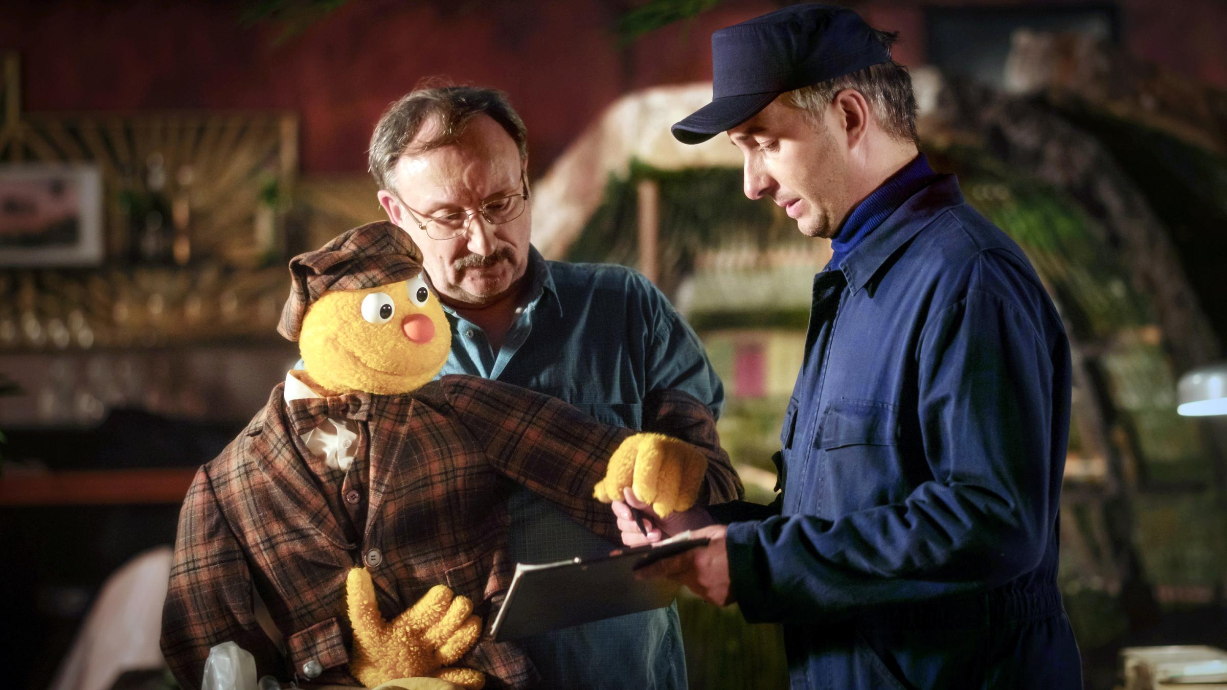 "Hallo Spencer - Der Film:" Die Handpuppe Spencer und Puppenspieler Jakob Sesam (Rainer Bock) unterschreiben einen Lieferschein, den ein Mann in blauer Arbeitskleidung (Jan Böhmermann) ihnen hinhält.
