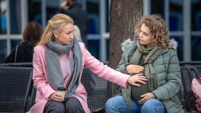 "Mein Kind - моя дитина": Judith Koch (Lisa Maria Potthoff) und Oksana Smirnova (Alina Danko) sitzen nebeneinander auf einer Bank. Judith hat eine Hand auf den Babybauch von Oksana gelegt. Die beiden schauen sich tief in die Augen.
