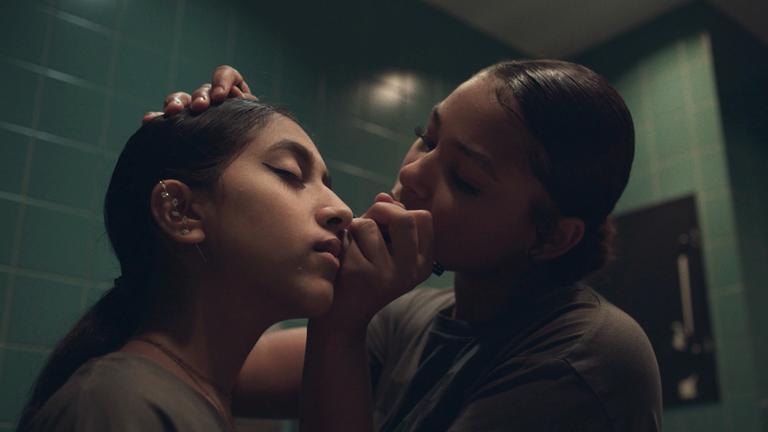 "Sisterqueens":  Filmfest München 2024: Die zwei Freundinnen Rachel (11) und Faseeha (12) stehen sich in einem gekachelten, halbdunklen Raum mit geschlossenen Augen gegenüber. Faseeha legt ihre rechte Hand auf Rachels Kopf; ihre linke Hand berührt deren Gesicht