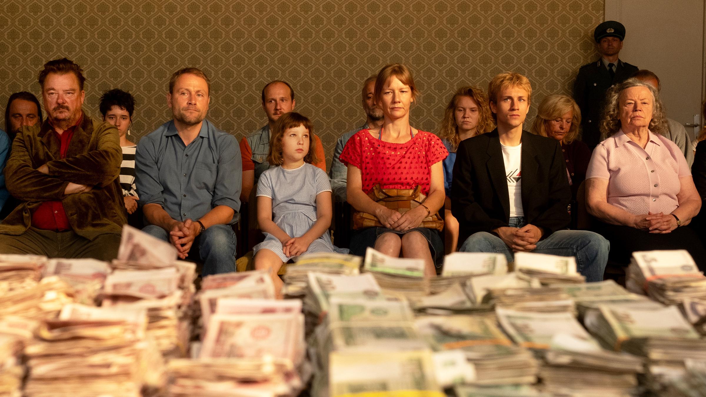 "Zwei zu Eins": Markowski (Peter Kurth), Robert (Max Riemelt), Dini (Lotte Shirin Keiling), Maren (Sandra Hüller), Jannek (Anselm Haderer), Käthe (Ursula Werner) sitzen mit mehreren Personen in zwei Stuhlreihen. Vor ihnen auf einem Tisch gestapelte Geldbündel.