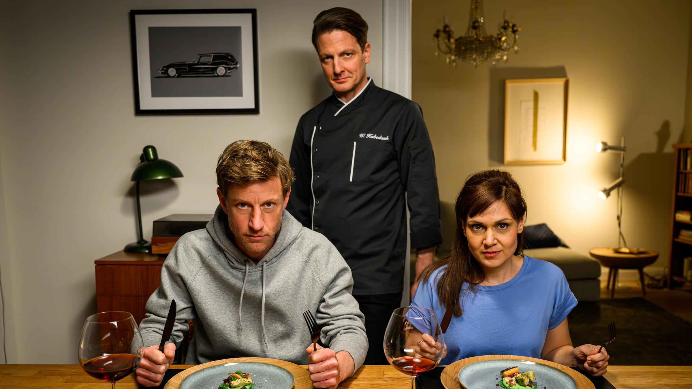 "Friesland – Sterneduell":  Henk Cassens (Maxim Mehmet) und Süher Özlügül (Sophie Dal) sitzen nebeneinander am Tisch. Vor ihnen stehen zwei angerichtete Teller mit Essen. Hinter ihnen steht Wolfgang Habedank (Holger Stockhaus) in einer Kochuniform.