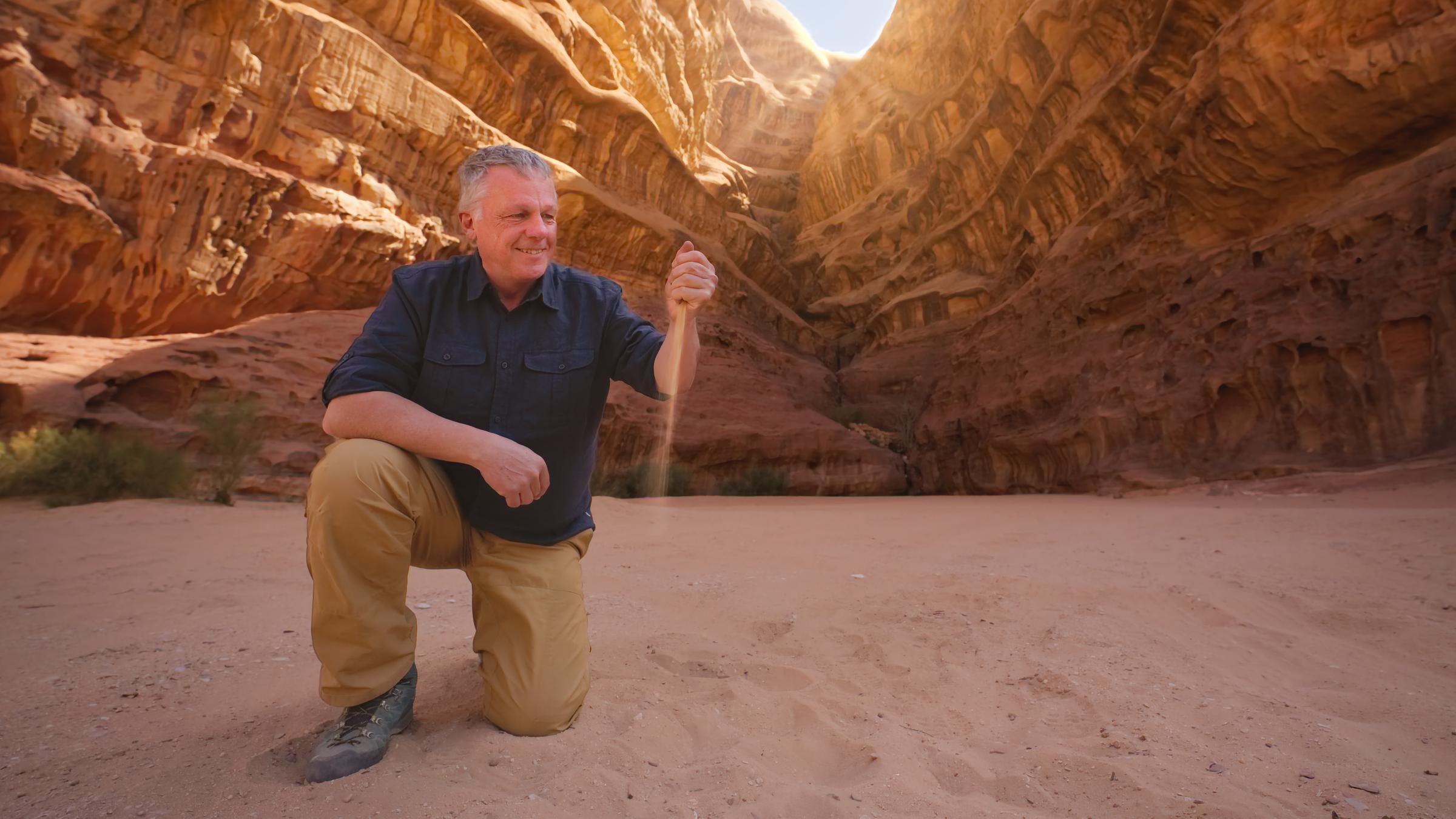 "Terra X: Der Metall-Planet - Wie Rohstoffe die Zukunft sichern": Colin Devey kniet in der jordanischen Wüste vor einer Felsformation im Sand und lässt Sand durch seine Faust rieseln.