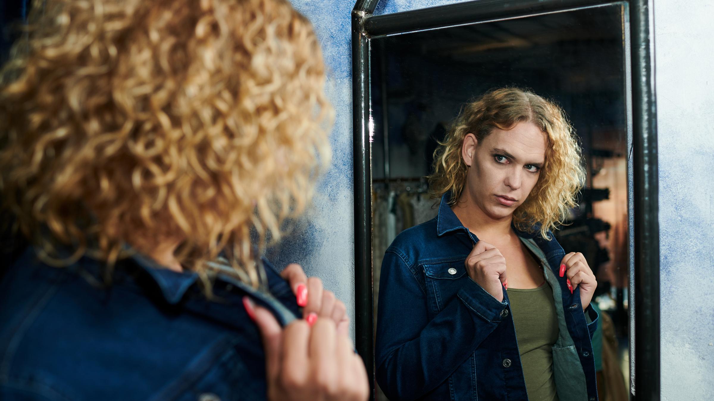 "37 Grad: Trans - Drei Generationen, eine Reise": Eine Frau mit blonden, lockigen, schulterlangen Haaren steht vor einem Spiegel und schaut hinein. Sie trägt eine Jeansjacke, darunter ein dunkelgrünes T-Shirt.