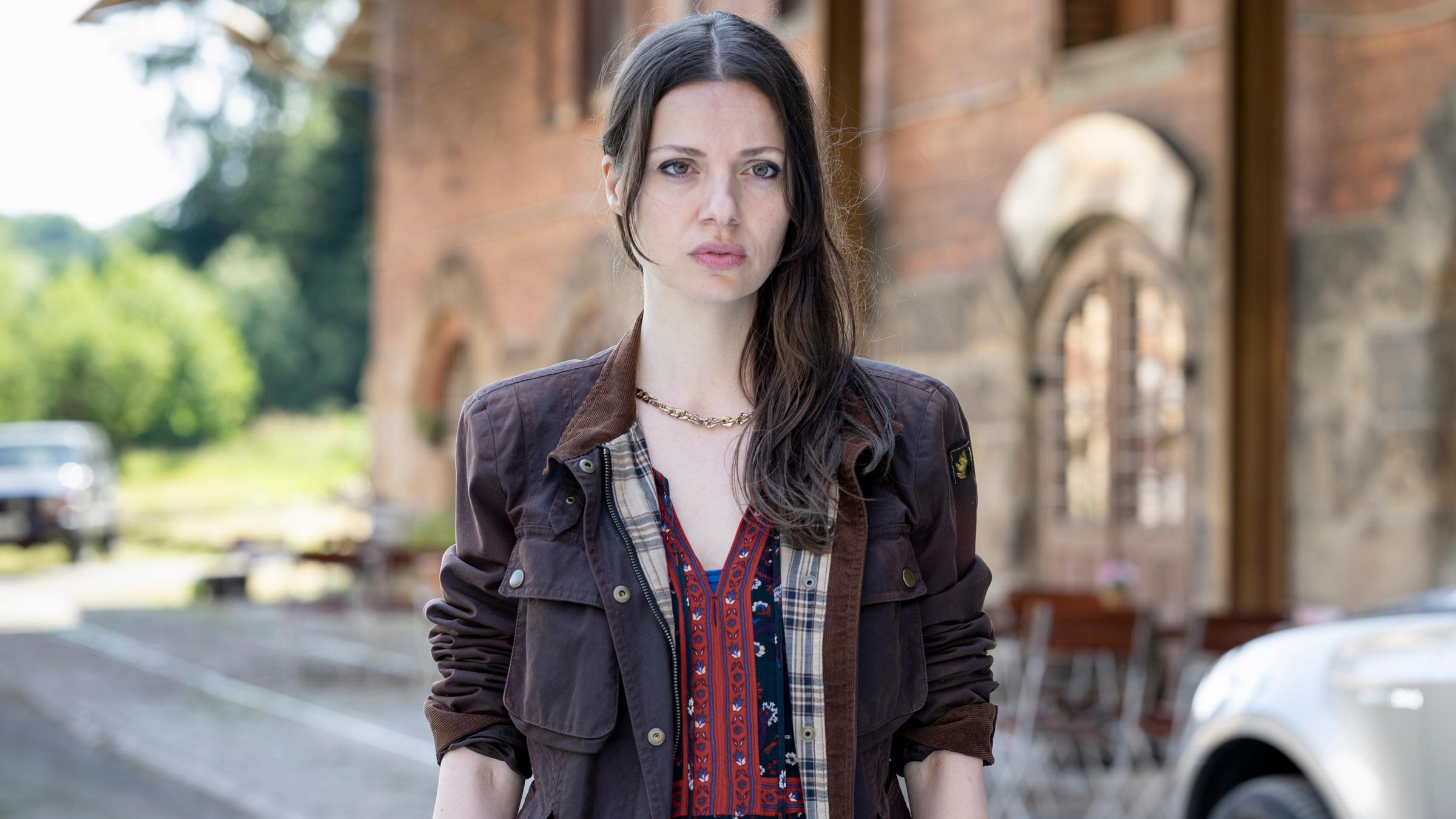 "Mandat für Mai - Vogtland": Mai (Julia Hartmann) steht vor der Dorfkneipe und blickt angespannt zur Seite. Im Hintergrund steht der Backsteinbau mit der Dorfkneipe, davor stehen Klappstühle und ein Auto.