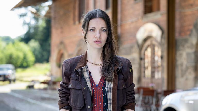 "Mandat für Mai - Vogtland": Mai (Julia Hartmann) steht vor der Dorfkneipe und blickt angespannt zur Seite. Im Hintergrund steht der Backsteinbau mit der Dorfkneipe, davor stehen Klappstühle und ein Auto.