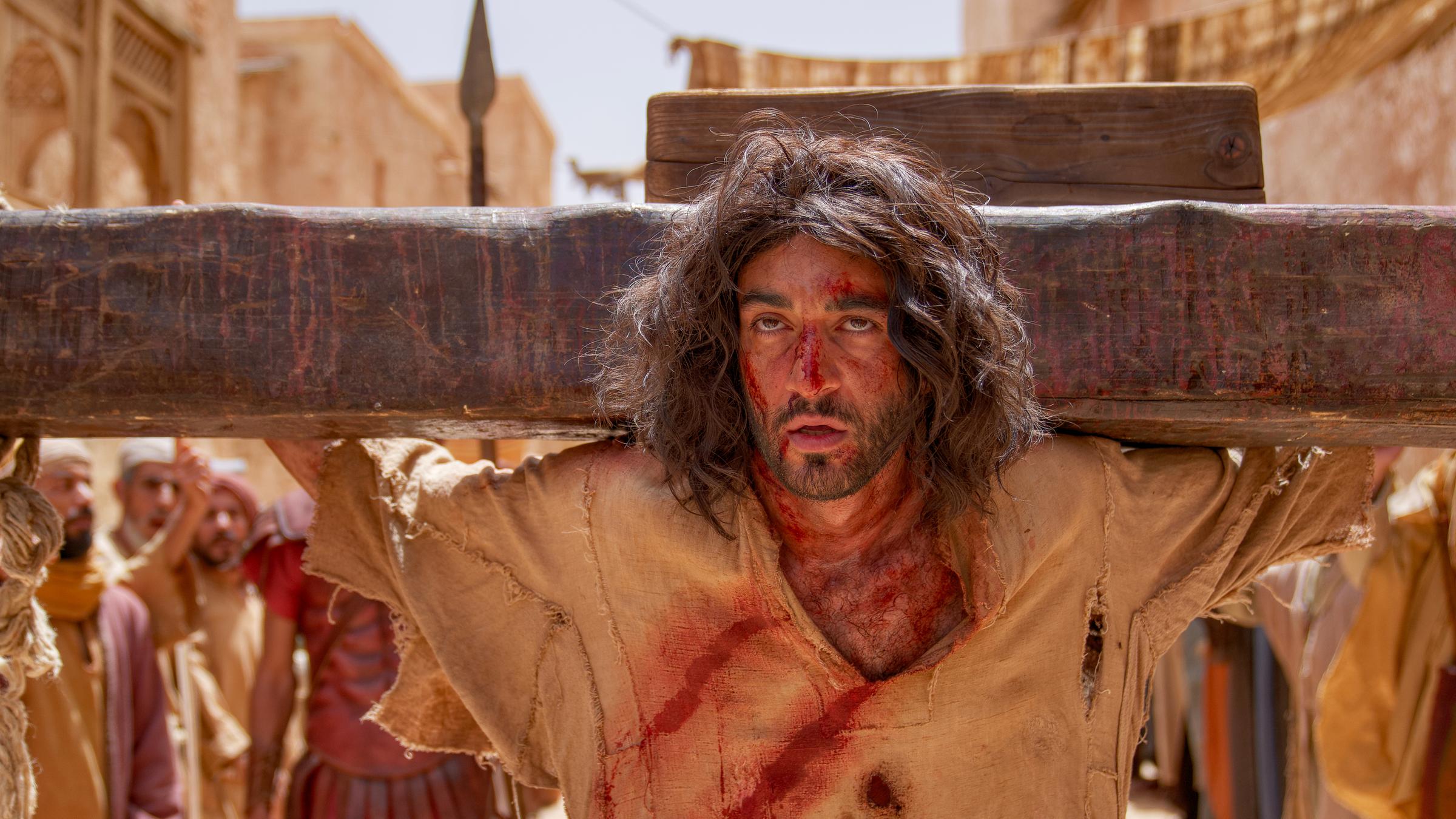 "INRI - Warum musste Jesus sterben?": Das Gesicht von Jesus (Eidin Jalali)  ist von Blutspuren gezeichnet. Dahinter liegt der Querbalken des Kreuzes. Im Hintergrund stehen Menschen.