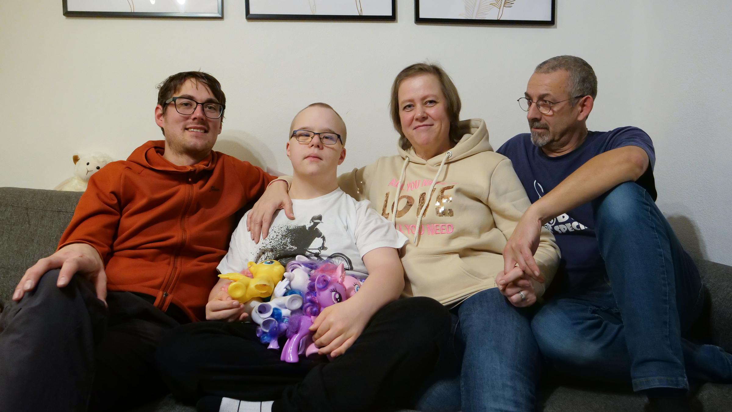 "Leben ist mehr! - Sterbebegleiter entdecken eine andere Welt": Tobias, ein Mann Mitte Dreißig, sitzt mit Jannes, einem Jungen mit Down-Syndrom, Yvonne, Jannes Mutter und deren Lebenspartner auf einer grauen Sitzecke. Jannes hat Einhörner auf dem Schoss.