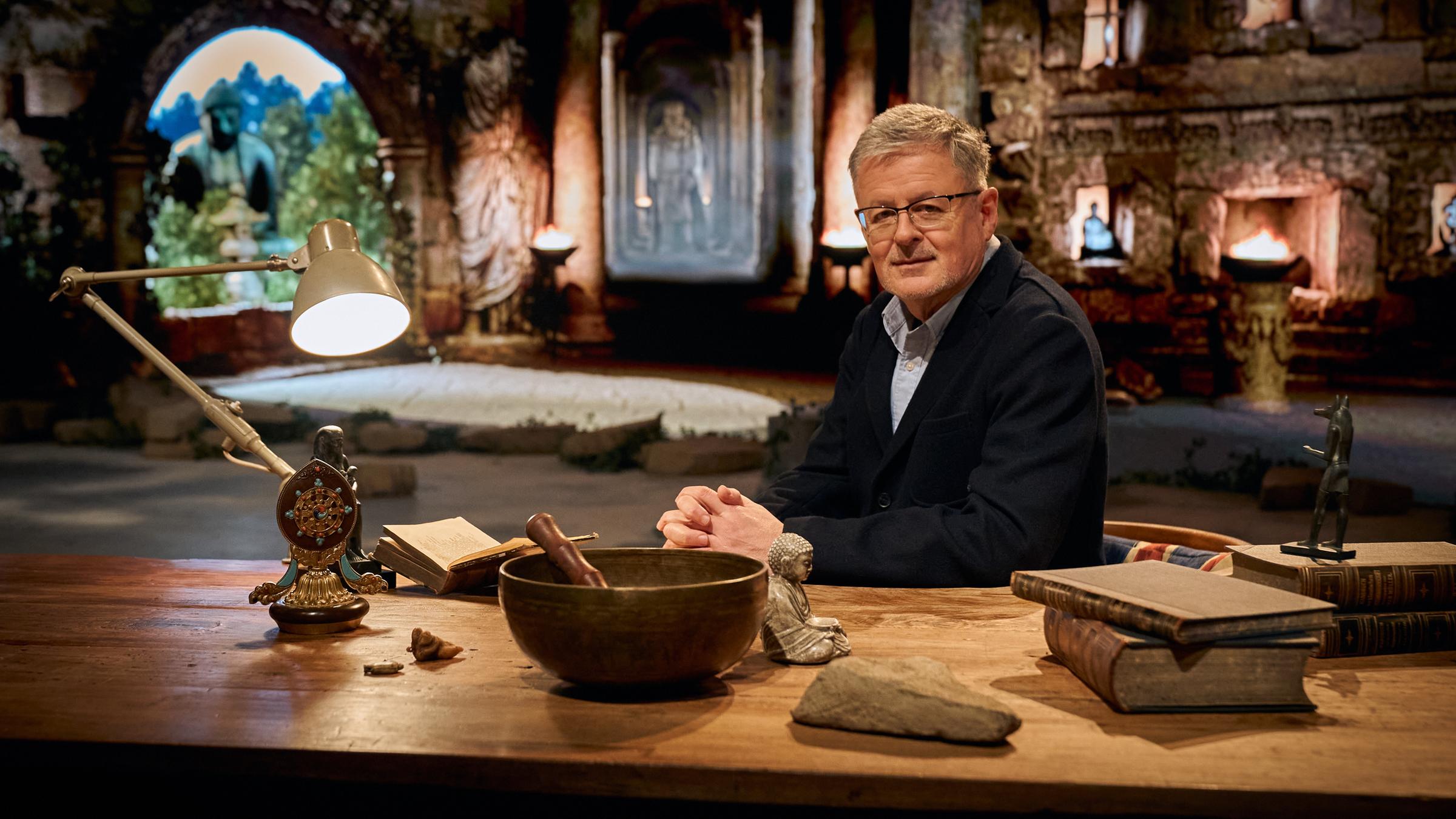 "Terra X: Macht der Götter - Weltgeschichte der Religionen - Der göttliche Funke - Wie entstand der Glaube?": Historiker und Moderator Christopher Clark sitzt am Schreibtisch eines Studierzimmers mit verschiedensten Merkmalen unterschiedlicher Religionsrichtungen.