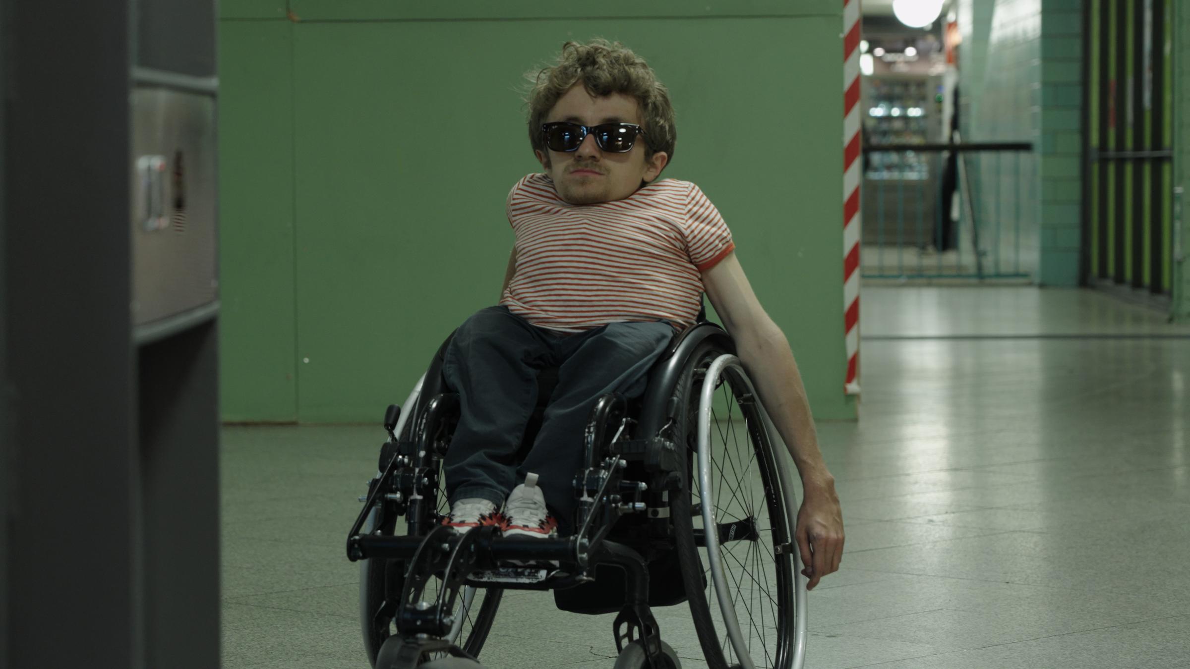 "Ab 18! – Die Selbstverständlichkeit des Seins": Ein kleinwüchsiger junger Mann mit rotgestreiftem T-Shirt und großer Sonnenbrille sitzt in seinem Rollstuhl und wartet auf den Aufzug.