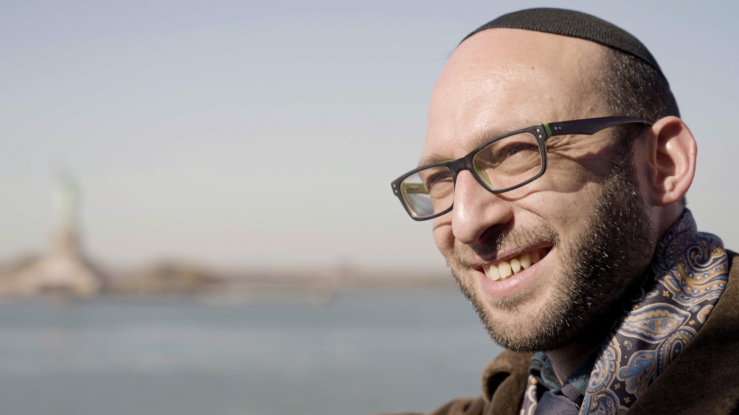 "Ultraorthodox: Der Kampf des Rabbi Akiva": Im Vordergrund ist Rabbi Akiva Weingarten im Portrait zu sehen, im Hintergrund ist die Freiheitsstatue sowie der Hudson River in New York in Unschärfe zu erkennen.