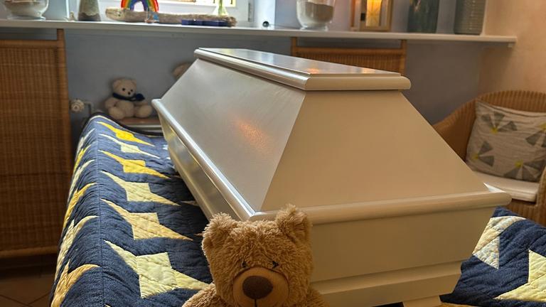 "Tabu Tod (1/2) - Sterben heute": Heller Kindersarg auf einer Sternendecke, davor ein Teddy, im Hintergrund ein Buntglasfenster und brennende Windlichter.